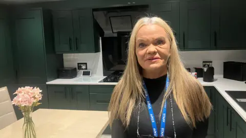 BBC Photo of Gaynor Hutton in the kitchen of her home in Collyhurst. She has long blonde hair and wears a black long-sleeved top.