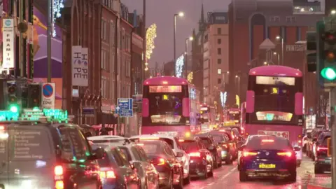 BBC A long line of cars and buses are at a standstill in Belfast city centre. it is evening and all the brake lights on the vehicles are on.