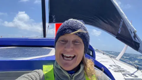 Pip Hare Ocean Racing Pip Hare is wearing a blue hat and has long blonde hair. She is smiling. Behind her is a black sail and white hull - the ocean is behind the boat.