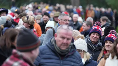 PA Media Members of the public gather ahead of the Christmas Day morning church service attended by the royal family at St Mary Magdalene Church in Sandringham, Norfolk. People are wearing puffer jackets, beanies and chat among themselves. 