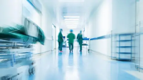 Audit Scotland Stock image shows a blurred hospital ward. In the distance you can see the figures of three people wearing scrubs and hats.