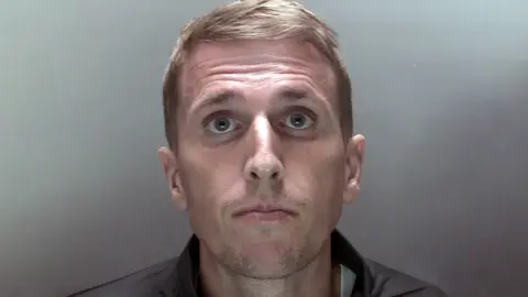 NCA Close up custody photo of Colin Wright - fair haired man with green eyes looking into the camera against a tan coloured background