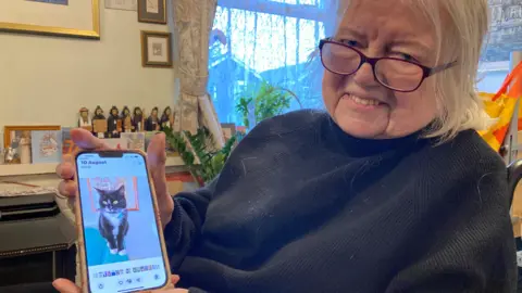 BBC A woman with mid-length white hair and glasses, wearing a black jumper, shows a photograph on her phone of a black cat with white paws and a white chest.