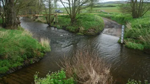 Geograph/John Walton A rural road crosses a river via a deep ford. There is a footbridge to the left, bushes and vegetation, and a white measuring post.