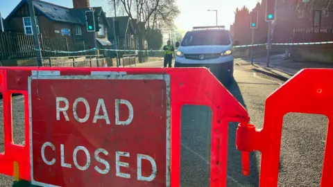 BBC The scene of the investigation where a 'road closed' sign can be seen in front of a police van and officer. There is police tape cordoning off the road in the background.