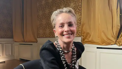 BBC Sharon Stone smiling at the camera wearing a black dress and silver beaded necklace