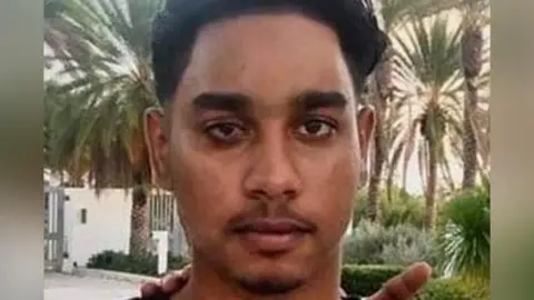 Family Handout A close-up of a young man's face. He has dark hair and eyebrows and a sparse moustache and goatee beard. He is outside, surrounded by palm trees and low shrubs. A white wall and gates can be seen in the background.