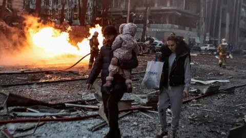 Reuters A man holding a baby walking away with flames and debris on a road 