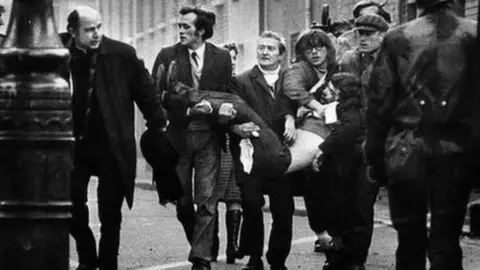 BBC An iconic image of Bloody Sunday in which a group of men carry one of the injured through the streets. A priest walks ahead and  waves a white handkerchief