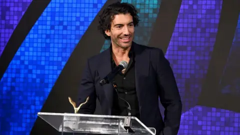 Getty Images Actor Justin Baldoni is seen on a stage accepting an award in New York. He is wearing a dark-coloured suit and is standing at a microphone with a golden award sitting on the lectern. 