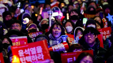 Reuters A close up of a sea of faces at a protest in South Korea. Several people are carrying signs or waving candles and flashlights.