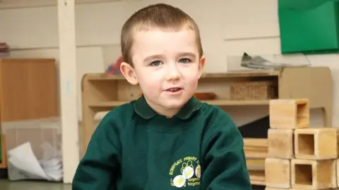 Essex Police Lincoln Button wearing his school uniform, which is a green sweater that has a green polo top underneath. He has short brown hair and is smiling at the camera while sitting in a classroom.