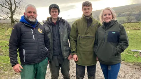 BBC Family (left to right) Matthew Staley, Lewis Staley, Luke Staley, Liz Staley. They are standing in a field. Matthew has a grey beard and a black jacket, Lewis is wearing a black jacket and a hat, Luke is wearing a green jacket and Liz is wearing a black jumper.