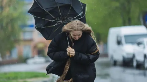 Getty Images A blonde woman stands under an umbrella which has turned inside out. She's in a black duvet coat and her face is in a grimmace. It's raining.

