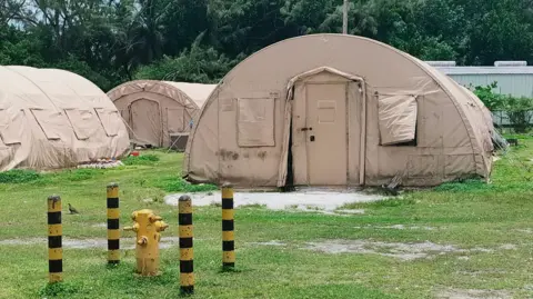 BBC Photo shows three round beige tents on green grass. In the background there is a hut