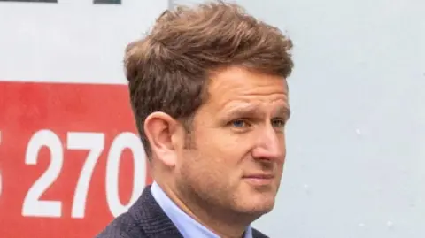 SWNS A man with brown hair, wearing a dark suit, looking away from camera