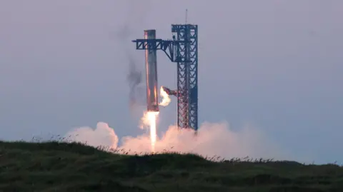 Reuters Starship's booster lowers itself into its launch tower as its jets power down. Fire is seen coming out of the bottom of the booster and smoke fills the surrounding air.