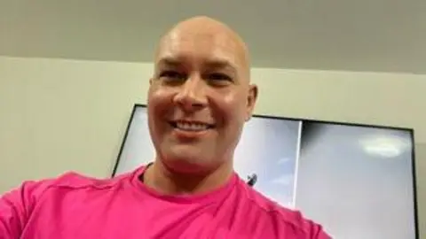 Family photograph Kirk Marsden, who is bald, smiles posing for a photograph in a pink top. 