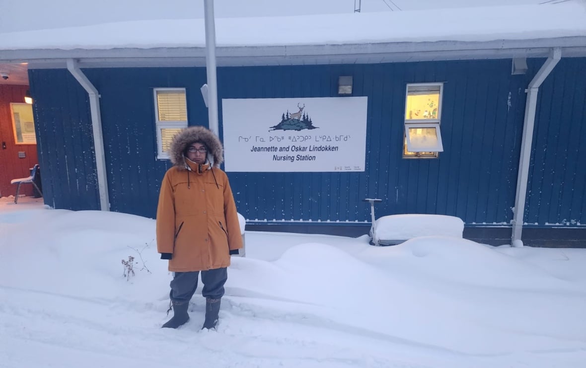 A person stands in the snow outside of a blue-coloured building.