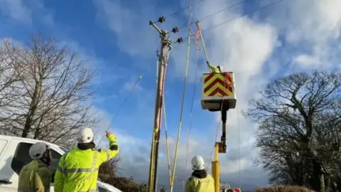 BBC Three workers in high vis clothing stand beside a white van parked next to a telegraph poll, which has a cherry picker beside it. Another worker is reaching up towards the wires.