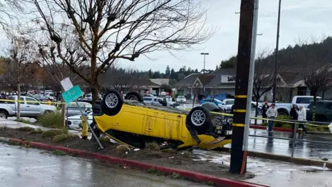 Scotts Valley Police Department A yellow five-door car that has been flipped upside down and onto the pavement by a tornado in Scotts Valley. A shop and a large car park with other parked cars can be seen in the background.