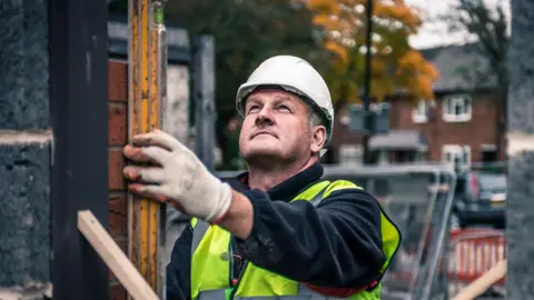 Getty Images A bricklayer wearing a white hard hat checks brickwork with a spirit level on a building site