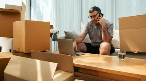 Getty Images Man sits on a sofa, talking on a mobile phone, with a laptop on a table in front of him and surrounded by boxes. 