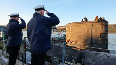 Royal Navy Two men salute as the submarine leaves the naval base. Men standing on the submarine in the distance can be seen to return the salute.