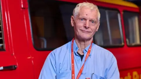 PA Media Royal Mail postman Robert Hudson looks towards the camera with a postvan in the background. robert is wearing a blue shirt with a red royal mail lanyard around his neck