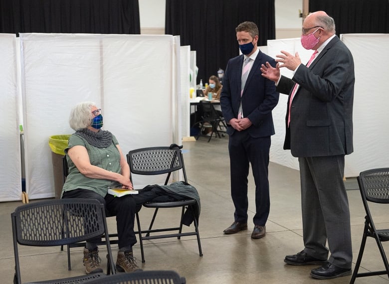 Two men in suits speak with a seated woman who has just received a COVID-19 vaccine.