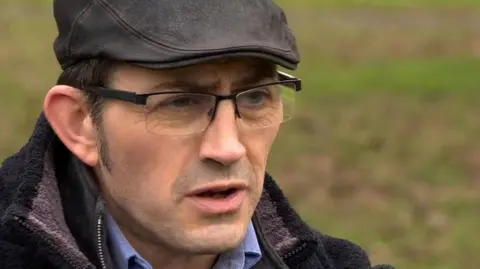 BBC Sean Faloon speaking to the BBC in 2018.  He has short, dark hair and is wearing thin-framed glasses, a black flat cap, a dark fleece and a blue shirt. 