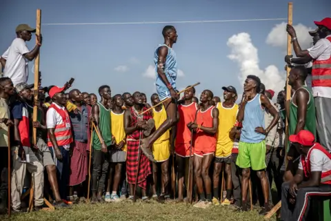 Luis Tato/AFP A Maasai athlete leaps vertically while taking part in the men's Maasai jumping competition, watched at close quarters by a number of spectators, some of whom are holding the high-jump frame.