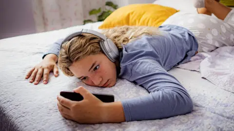 Getty Images A young teenager lies on a bed with a phone in her hand and is wearing headphones.