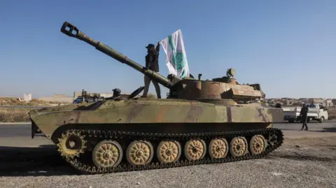 Reuters A Syrian rebel fighters waves the flag of Hayat Tahrir al-Sham (HTS) on top of a tank in Saraqeb, Idlib province, Syria (1 December 2024)