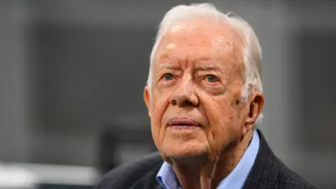 Getty Images Jimmy Carter, an elderly, wrinkled man with white hair. He is wearing a blue-collared shirt with a black, checkered sports jacket