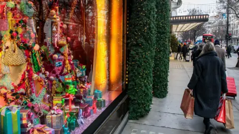 Getty Images Christmas shoppers in London. A window display is packed with Christmas trees and decorative presents wrapped in shiny paper and tied with ribbons. Outside, a shopper dressed in a warm coat carries shopping bags. 