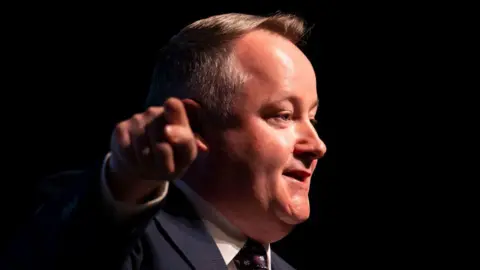 Getty Images Darren Millar is pointing towards the camera while looking away at a right angle towards an audience. He is wearing a suit, tie and shirt, and is stood before a dark background.