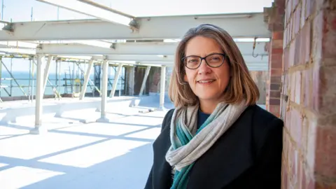 Hastings Commons Dr Jess Steele - wearing glasses, a black coat as well as a blue and white scarf - stands on the roof a building. Steel girders and scaffolding can be seen behind her with the sea observable in the distance.