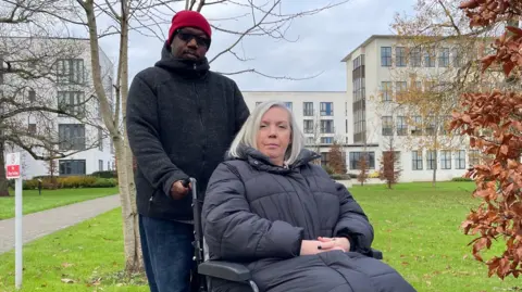 Ben Schofield/BBC Karen James, seated in a wheelchair and looking direct to camera, with her husband Mansfield holding the chair's handles. They are outside and Karen is wearing a thick black coat that ends below her knees. She has silvery hair cut into a bob. Mansfield is standing and wearing a red bobble hat, dark glasses, a dark fleece and blue jeans. They are on the edge of a grassed area, with white art deco style buildings and some trees in the background. 