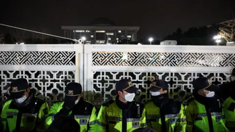 Reuters Police in caps, facemasks and high-viz jackets stand guard in front of the gate of the National Assembly after South Korea's president declared martial law