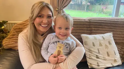 BBC A picture of a blonde woman wearing a light pink top sat on a brown sofa, cuddling her two-year-old son who has blonde hair and blue eyes, smiling at the camera.