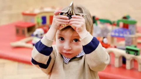 Handout George wearing a beige jumper and white and blue stripe shirt. He holds a star award above his head. In the background is a blurred playmat and wooden toys.