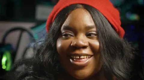 GeekyCassie A woman with long black hair and wearing a red hat smiles while looking away from the camera