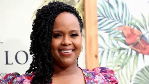 Natasha Rothwell is smiling, with only her head visible. She is wearing a pink floral dress and has her hair swept to one side. There is a floral White Lotus themed backdrop behind her