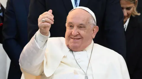 EPA Pope Francis gestures and smiles to the crowds during a papal visit to Corsica in December 2024. He is wearing white robes, a white cap and a crucifix.