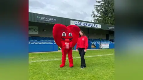 Faith Harrison Faith has long brown hair and is standing on the grass of a football pitch wearing a red British Heart Foundation hoodie. She is standing next to a person that is dressed in a full-body red heart costume with a British Heart Foundation logo on it.