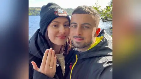 Faith Harrison Faith is wearing a black coat and dark coloured beanie, and is holding up her hand. On her finger is an engagement ring. Next to her is her partner Sam, who has short dark brown hair and black facial hair. Behind them is a lake.