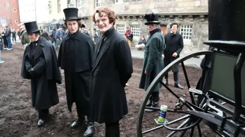 PA Media Actors on the set of Frankenstein. Three men are smiling at the camera while dressed in 18th century coats and shoes, with two of the men wearing hats. Other extras mill about in the background. 

The street is all mud and earth. 