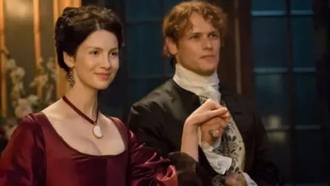 STARZ/AMAZON PRIME Caitríona Balfe and Sam Heughan in a scene from Outlander - she is dressed in a red dress and has dark hair, while he is dressed formally. They are holding hands and smiling, and appear to be about to take to the dance floor.