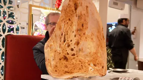 Reporter David Sillito almost entirely obscured by a giant hanging naan bread in a restaurant